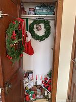 Photo of closet door hung with two faux greenery wreaths decorated with pinecones, red berries, ceramic bird ornament, and red bead garland, and a red Christmas stocking. Shelving inside closet shows assorted holiday tins, candles, poinsettia decorations, and a ceramic teapot.