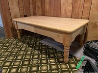 Angled side view of the wooden coffee table showing one turned leg and rounded corner detail on wood grain tabletop.