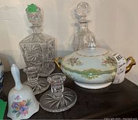 Two pinwheel cut glass decanters, two cut glass candle holders, Noritake soup tureen with lid, and ceramic bell shown together on shelf.