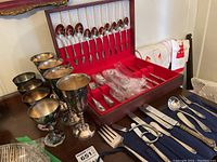 Wooden box open showing stainless steel cutlery arranged inside and tall, tarnished silver plate goblets alongside it