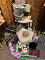 Photo of lot showing white shower chair, portable bedside commode buckets stacked on it, beige elevated toilet seat wrapped in plastic, purple slip-on slippers on floor, and various small medical items on a tray including a medical diary and baby monitor.