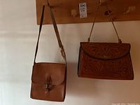 First photo showing both leather purses hanging on a wall with good lighting. Left purse is smaller, plain leather. Right purse is larger with detailed tooling.