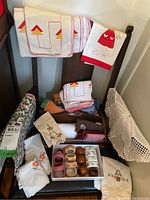Photo showing assorted embroidered linens including doilies, napkins, kitchen towels, placemats, tablecloth, and napkin rings arranged on and leaning against a chair.
