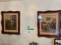Two ornate gold-tone wood framed prints with maroon mats and glass fronts hanging on a wall, showing vintage-style scenes with people and animals.