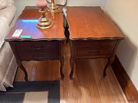 Two matching wooden side tables with curved rectangular tops and two drawers each, placed on a wood floor next to a couch.