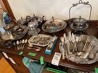Photo of various silver plated serveware items including teapots, coffee pot, trays, and cutlery on a wooden surface.