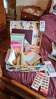 Wide view of a floral box on a pink quilted surface filled with assorted greeting cards and several sheets of postage stamps placed outside the box.