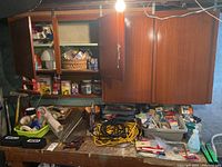 Wide view of wooden wall cabinet open with shelves containing hardware cans and containers, workbench below filled with hand tools, extension cords, and hardware components scattered on the surface.
