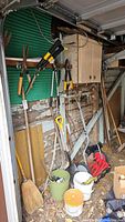 Wide view of garden tools against garage wall showing pruning shears, rakes, push mower, buckets with tools, and broom