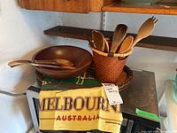 Photo of wooden kitchen utensils in woven basket container on serving tray with wooden salad bowl and dish towel nearby.