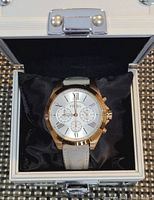 Fossil wristwatch with white dial, rose gold bezel, and beige leather strap placed on black cushion inside white metal box with lock.
