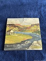 Front view of signed landscape oil painting on masonite showing river, cabin, trees, hills in fall colors, with visible crack at top.