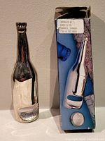 Bottle-shaped chrome metal bottle opener next to its original box labeled with importer and Canada location.