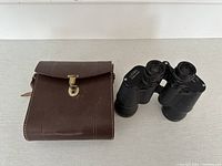 Lumar binoculars with brown carrying case resting beside them on a light surface.