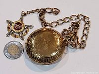 Engraved gold-tone hunter case pocket watch with attached chain and enamel fob seen alongside a Canadian toonie for scale