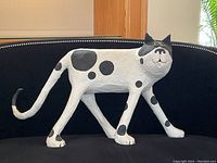 Three-quarter front view of the white and black spotted cat sculpture displayed on a black sofa