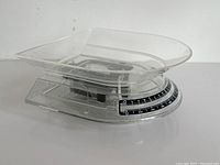 Side angle showing the clear plastic body, the rectangular weigh tray on top, and the mechanical dial on the front with weight markings in black and white.