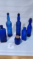 Full front view of the six cobalt blue bottles arranged on white fabric background showing their relative sizes and shapes