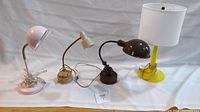 Photo showing all four lamps from left to right: pink adjustable arm desk lamp with clamp, cream adjustable arm desk lamp with round base, dark brown non-adjustable desk lamp with fixed arm, and yellow base table lamp with white fabric lampshade.