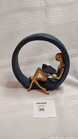 Side view showing the woman seated inside the circular frame reading a book, highlighting the dark blue speckled finish and gold coloring on the figure.