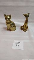 Two brass cat figurines side by side on a white surface with auction lot number card.