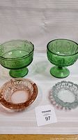 Four vintage glass items displayed on white cloth: rose pink ashtray, clear ashtray, green glass pedestal candy dish, and green glass dessert bowl.