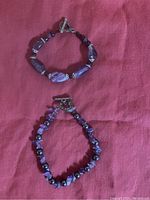 Photo showing two purple amethyst stone bracelets on a red fabric background, both with toggle clasps and silver spacer beads on one bracelet.
