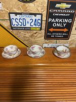 Three antique teacup and saucer sets displayed on a wooden shelf against a wall with vintage license plates and parking signs.