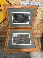Two framed artworks by Kim Norlien showing lakeside cabins in wood frames with green mats and artist nameplates.