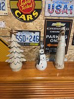 Three Christmas decorations arranged on a wooden shelf with vintage license plates and signs in the background, featuring a white layered tree, a glass snowman, and a tall Santa Claus figure.