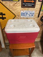 Photo of cooler closed, showing red body and white lid with four cup holders, placed on a wooden stool with background of road signs and license plates.