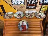 Five collectible porcelain plates by David Wright depicting indigenous female figures, displayed on wooden slats, background with license plates and signs