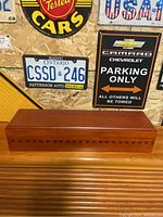 Closed rectangular wooden box placed on wood surface with license plates and signs in background, showing its polished exterior and wood grain.
