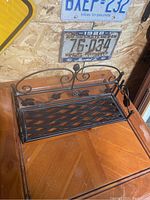 Front view of folding bronze metal decorative shelf sitting on wooden surface with license plates on wall behind. Shows entire shelf including grapevine ornamentation on backrest and lattice pattern on shelf surface.