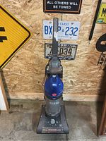 Front view of upright Hoover Windtunnel vacuum cleaner showing blue and gray body with clear dust bin, control buttons, and base with brush.