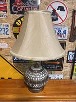 Table lamp with round patterned black and white base and beige fabric lampshade, photographed on a wooden surface against a background with license plates.