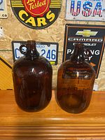 Two amber glass jugs positioned side by side on a wooden shelf with various vintage signs in the background.