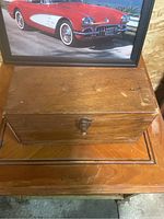 Closed rectangular wooden box with a metal latch on top of a wooden surface, framed car photo behind.