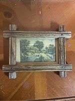 Top view of the framed artwork with visible rustic wooden frame and the image inside showing a mill by a water body and a dirt road.