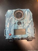 Front view of wild game trail camera showing lens, infrared LEDs, LCD screen, and control buttons with camouflage design.