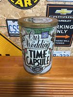 Closed metal tin labeled 'Our Wedding Time Capsule' placed on a wooden surface with decorative license plates on the wall behind.