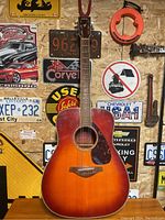 Full view of Yamaha acoustic guitar sunburst finish leaning against wall with various signs in background.