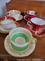 Photo showing four vintage bone china teacups and matching saucers from above and side angles, displaying intricate floral and gold designs in various colors including green, red, and pink.
