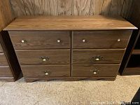 Front view of wooden dresser with six drawers, metal and ivory knobs, visible wood grain.
