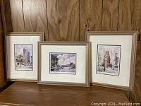 Three framed watercolor paintings by Blaine Jackson featuring Toronto landmarks, shown together on a shelf against a wood wall.