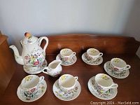 Full set displayed on wooden surface showing teapot, creamer, and five cup-and-saucer pairs with floral design.