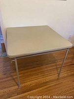 Front view of the square folding table set up with beige top and metal legs on wooden floor.