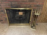 Full view of gold toned fireplace screen with attached black mesh and matching utensil set on stand to the side.