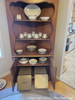 Open cabinet displaying cups, saucers, three stacks of plates and contributing serving pieces with boxed settings stored below.