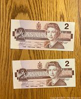 Front view of two Canadian $2 bills laid flat on a wooden surface, displaying the portrait of Queen Elizabeth II and denomination details.